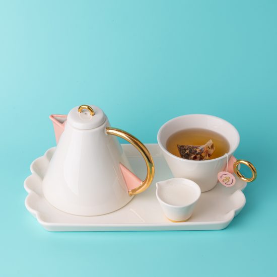 Une théière, une tasse blanche qui contient du thé et une petite coupelle blanche qui contient du lait, sur un plateau blanc - fond bleu clair